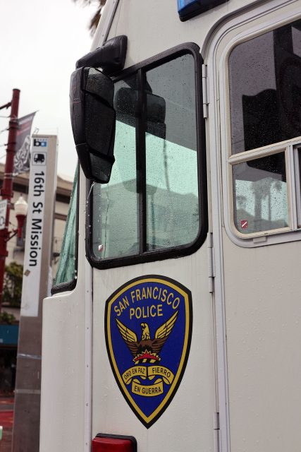 Side view of a San Francisco Police vehicle with a visible police badge, near a 16th Street Mission sign.