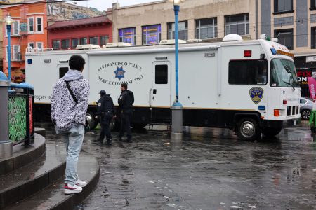 A person stands near a San Francisco Police mobile command unit parked on a wet street with officers nearby.
