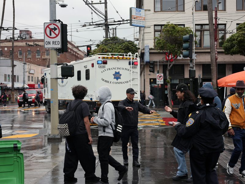 Group of people and police officers standing on a wet city street near a mobile command vehicle. A "No Turn On Red" sign is visible.