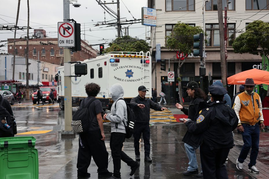 Group of people and police officers standing on a wet city street near a mobile command vehicle. A "No Turn On Red" sign is visible.