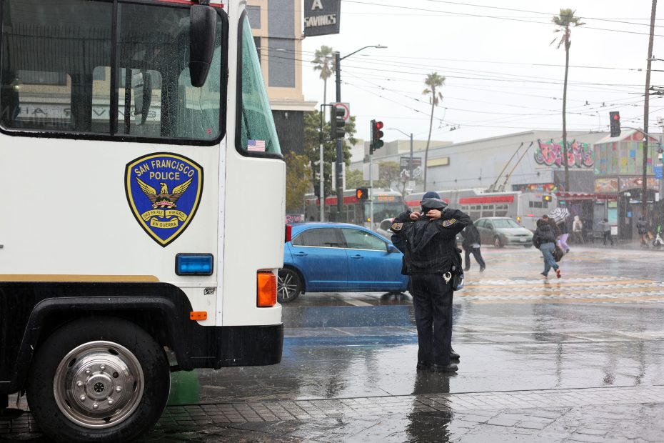 Police officer directing traffic in the rain near a San Francisco Police Department vehicle at an intersection.