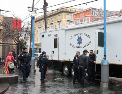 Police officers stand near a San Francisco Police mobile command vehicle on a rainy street, with pedestrians walking by.