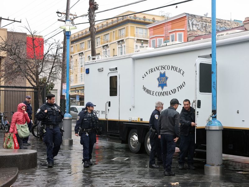 Police officers stand near a San Francisco Police mobile command vehicle on a rainy street, with pedestrians walking by.
