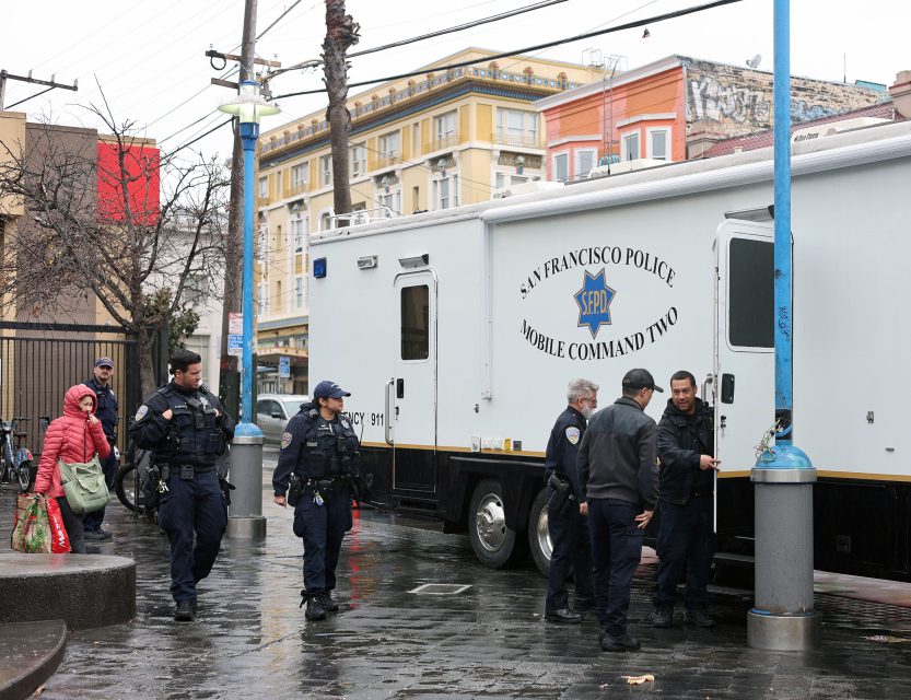 Police officers stand near a San Francisco Police mobile command vehicle on a rainy street, with pedestrians walking by.