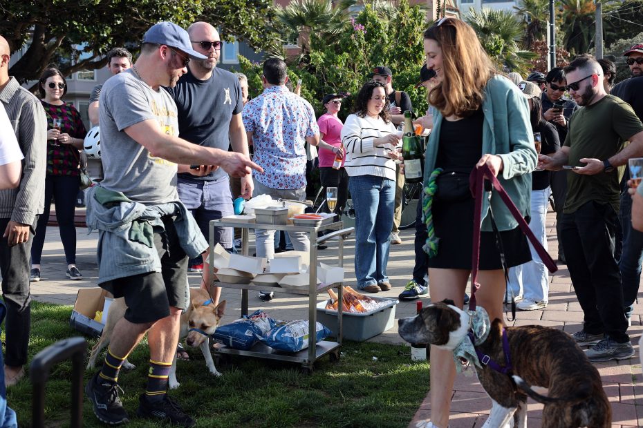 A group of people gathered outdoors, some interacting with dogs on leashes. A food cart with supplies is visible in the background.