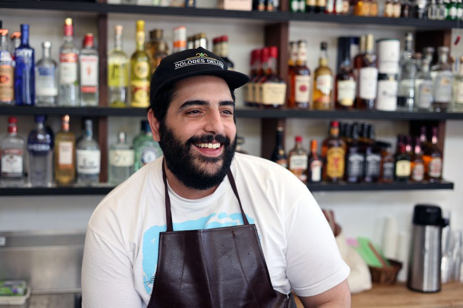 A bearded man wearing a cap and apron smiles while standing in front of a shelf filled with liquor bottles.