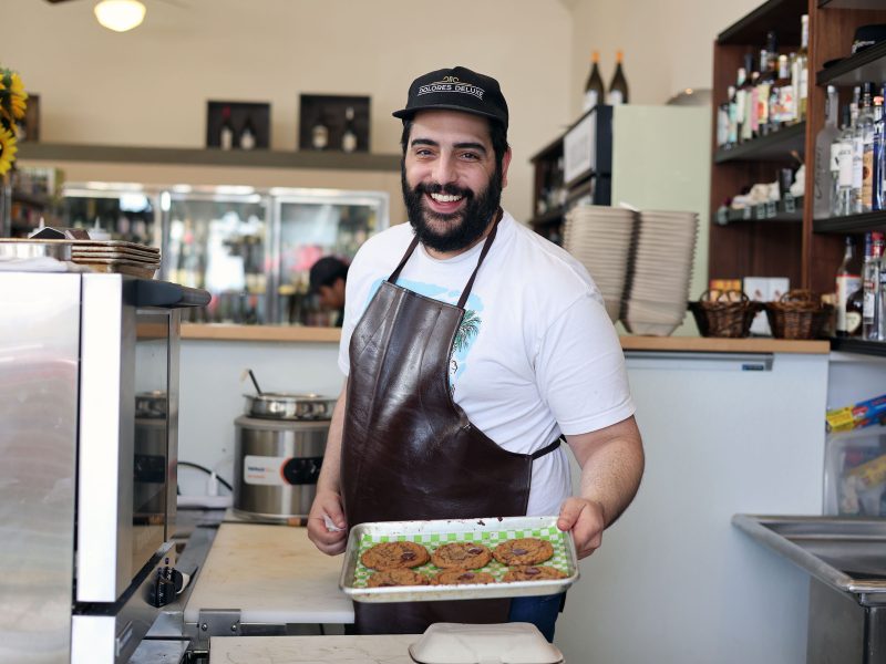 A man in an apron and cap holds a tray of cookies in a bakery setting.