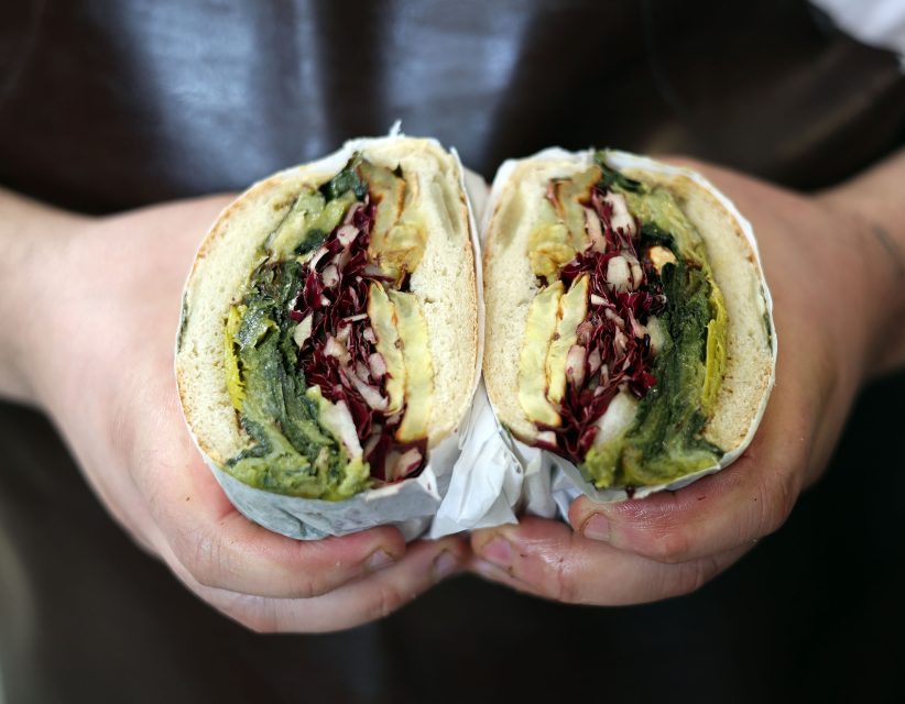 A person holds a sandwich cut in half, filled with layers of greens, red cabbage, and toppings, wrapped in paper.