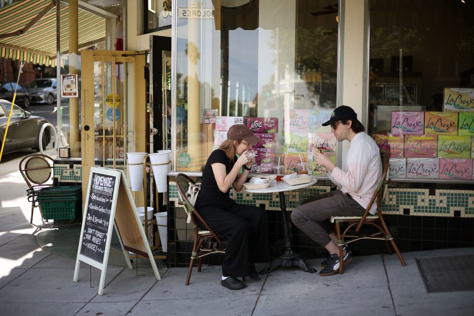 Two people sit at a small table outside a storefront, eating and drinking. A chalkboard menu and stacks of LaCroix boxes are visible in the background.