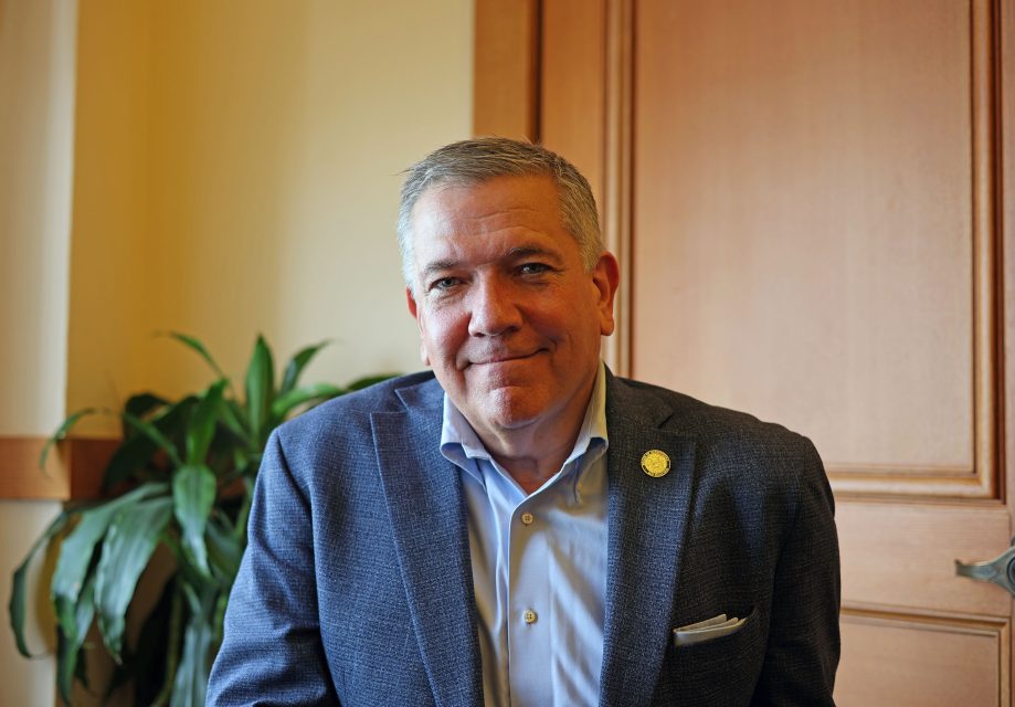 A man in a blue blazer and light shirt sits in front of a wooden door, with a potted plant visible beside him.