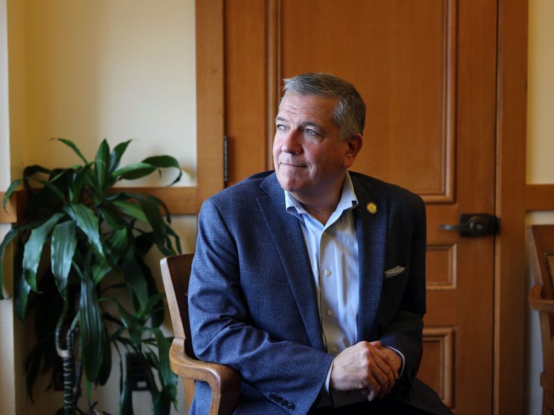 A man in a blue blazer sits on a chair in an office with a wooden door and a potted plant in the background.
