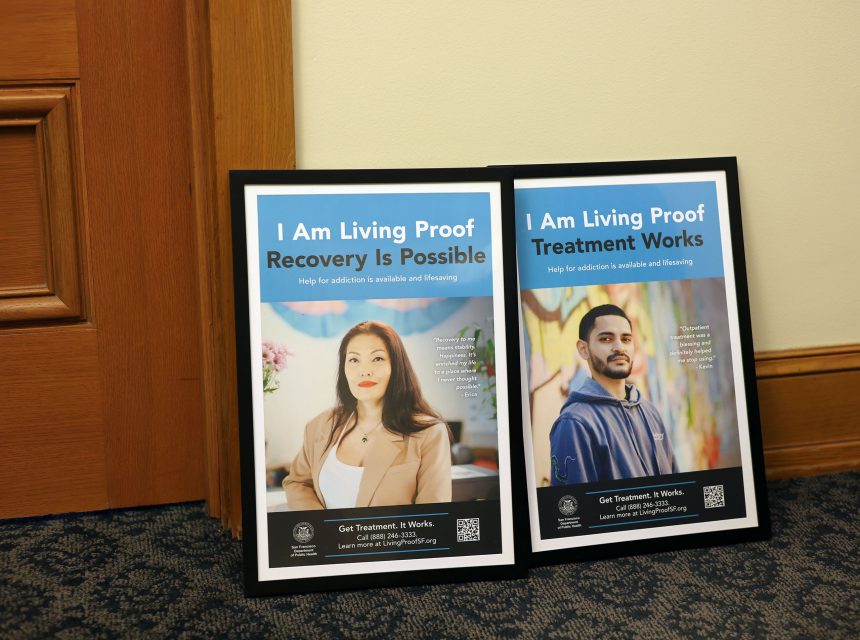 Two framed posters on the floor feature individuals with text promoting addiction recovery, stating "I Am Living Proof" with messages about treatment and recovery.