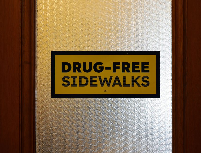 Sign on a frosted glass door reads "Drug-Free Sidewalks" in bold black letters against a yellow background.