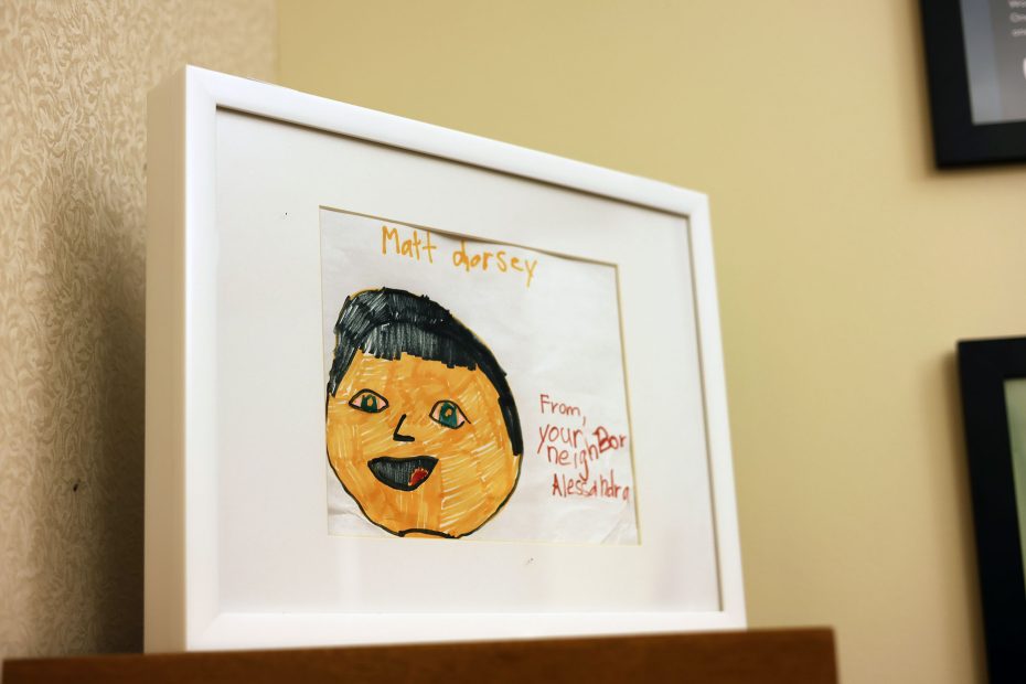 Framed child’s drawing of a person with "Matt Dorsey" written on top and "From your neighbor Alessandra" on the side, displayed on a wooden surface.