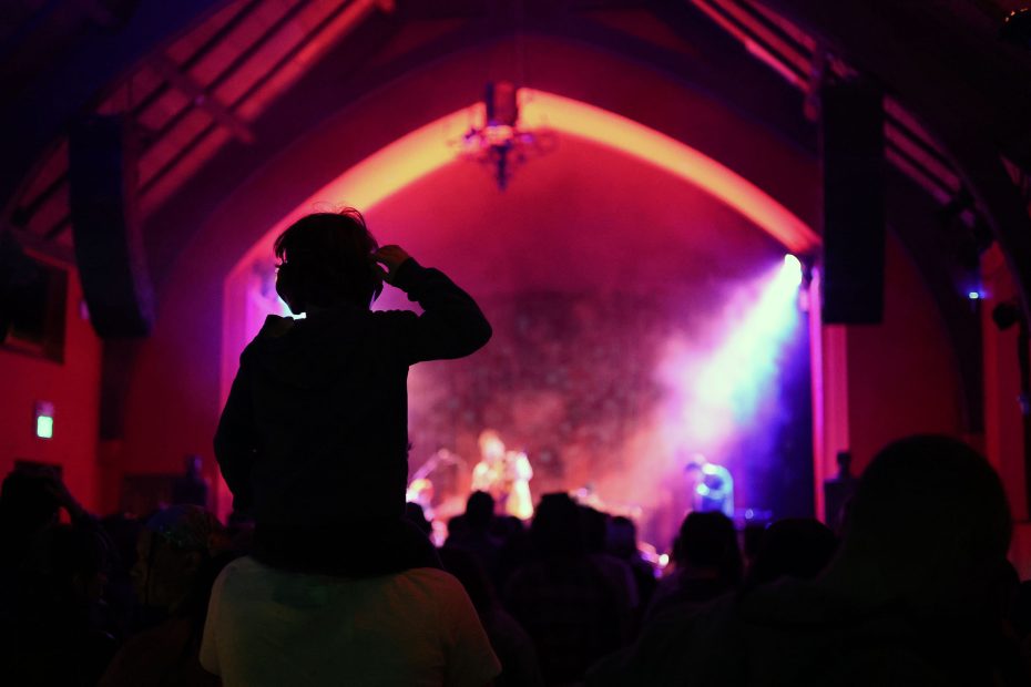 A child on someone's shoulders watches a live concert in a dimly lit venue with colorful stage lights.