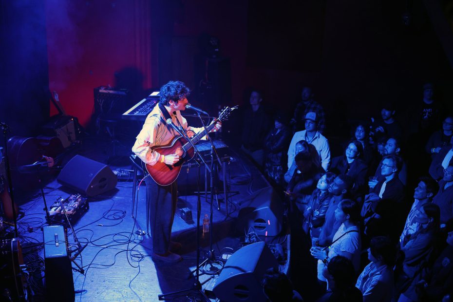 A musician plays an acoustic guitar on stage under blue and purple lights while a crowd watches attentively.