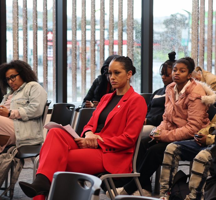 A woman in a red suit sits attentively holding papers in a room with others seated, some taking notes. The background shows large windows and a rainy scene outside.