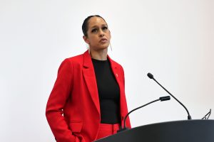 Person in a red suit speaks at a podium against a plain white background.