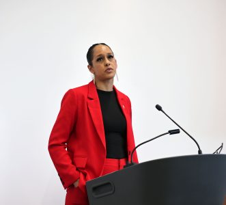 Person in a red suit speaks at a podium against a plain white background.