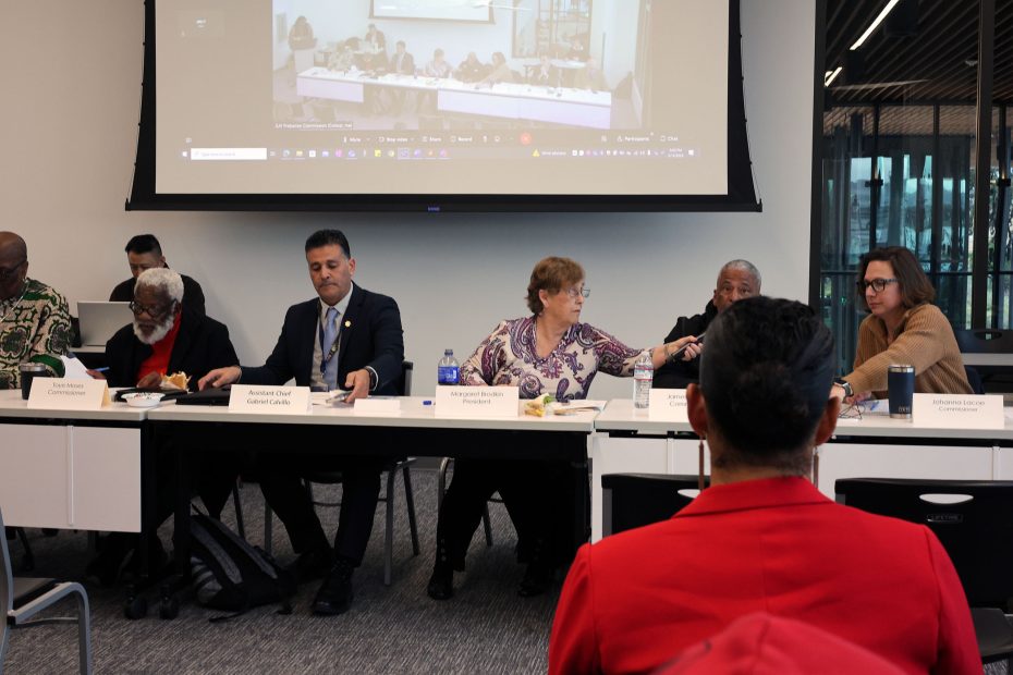 A group of individuals seated at a conference table in a meeting. A large screen displays a video call with additional participants in the background.