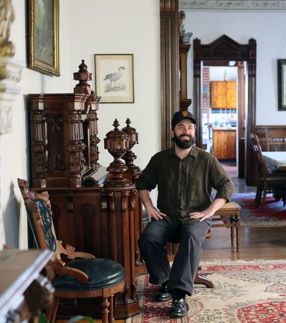 A person with a beard and cap sits on an ornate wooden chair in a vintage-style room, reminiscent of a classic film set, where intricate woodwork frames the scene and a dining area lingers in the background like a film camp backdrop.