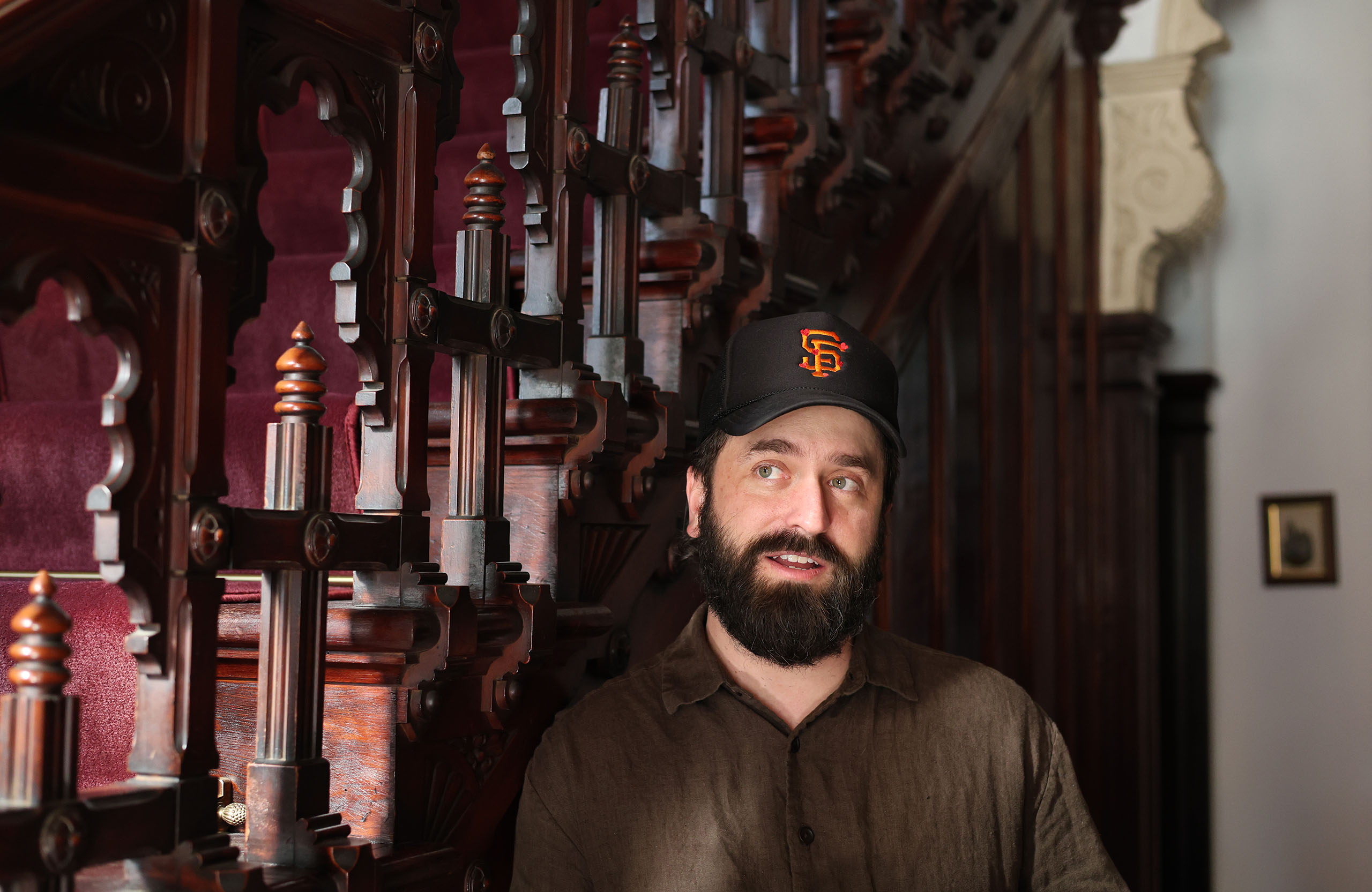 A man with a beard, wearing a dark cap and brown shirt, stands near an intricately carved wooden staircase with a red carpet, reminiscent of a classic film set.