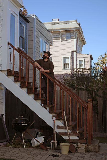 A man stands on an outdoor staircase, looking up as if awaiting a cue at a film camp. Gardening tools and a grill frame the scene, while two houses rest under the expansive blue sky.
