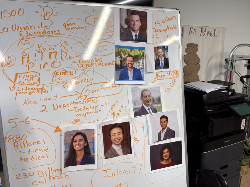 A whiteboard filled with handwritten notes in Spanish, formulas, and photos of six people attached to it. A photocopier is visible in the background.