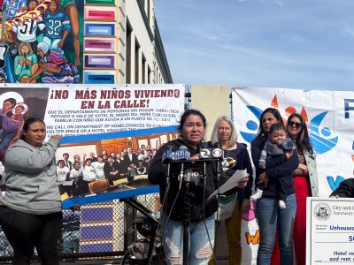 A woman speaks at a microphone during an outdoor event. People stand behind her near banners and a mural on a building wall.