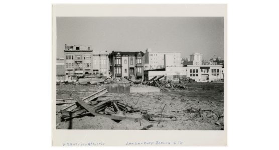 Demolished urban site with debris in the foreground, partially intact buildings mid-ground, and cityscape in the background.