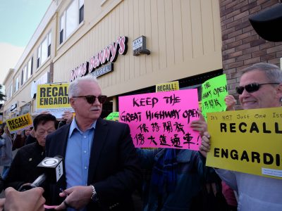 A group of people hold signs in a public protest outside a building, with messages about recalling "Engardio" and keeping the Great Highway open. A man in sunglasses stands in the foreground.