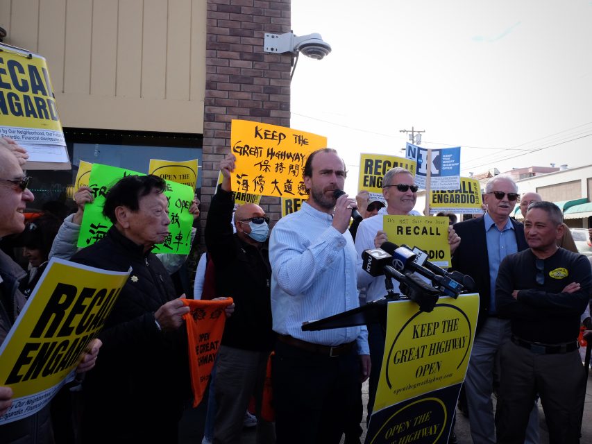 A man speaks at a podium during a protest. People around him hold signs advocating to "Recall Engardio" and "Keep the Great Highway Open.