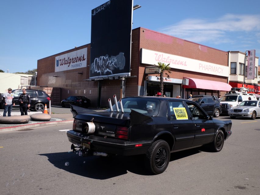 Black car with decorative exhaust pipes and a "Recall Newsom" sign on the rear window, parked near a Walgreens.
