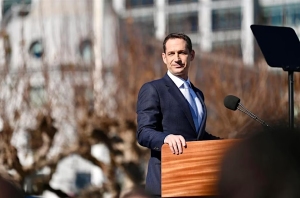 A person in a suit stands at a podium outdoors, speaking into multiple microphones. Trees and a building are visible in the background.