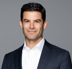 Man with dark hair in a suit and white shirt smiling against a gray background.