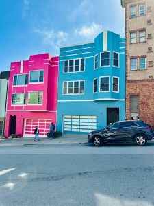 Two colorful houses in pink and blue stand side by side, each with contrasting white trim. A black car is parked on the street in front, with two people walking on the sidewalk nearby.