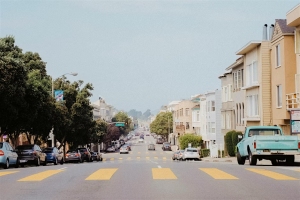 Street view with parked cars, trees, and residential buildings on both sides. Yellow pedestrian crossings are painted on the road. A vintage blue truck is parked to the right.