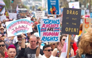 A diverse crowd marches, holding signs advocating for trans rights, including messages like "Trans Rights Are Human Rights" and "Protect Trans Kids.