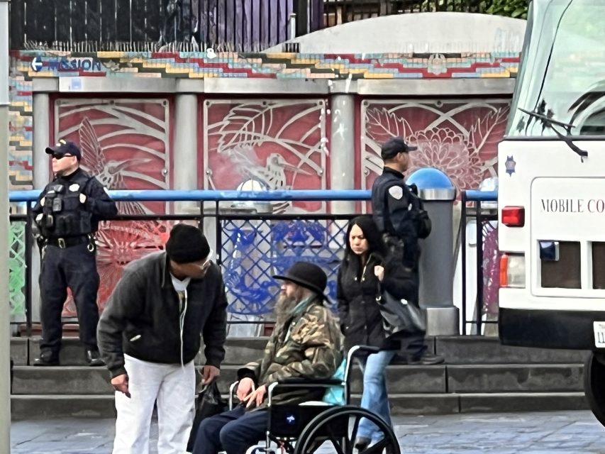 Two people converse beside a man in a wheelchair at the 16th St. plaza, near a mural with police officers standing in the background. A mobile command vehicle is parked close by.