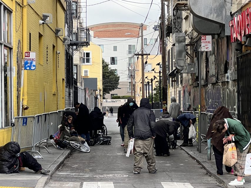 People gathered in a graffiti-lined urban alley, some sitting, others standing. A bicycle and shopping bags are visible. Urban buildings and "No Parking" signs are in the background.
