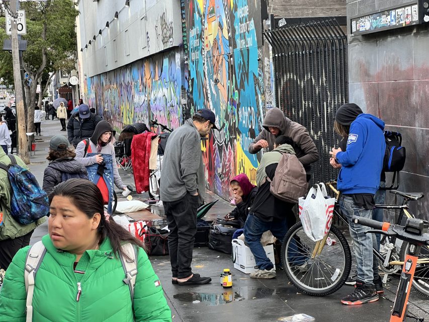 Crowds converge on the lively, graffiti-covered 16th St. plaza, with some seated and others standing. Scattered personal items and a bike embellish the scene as a woman in a green jacket strides past.