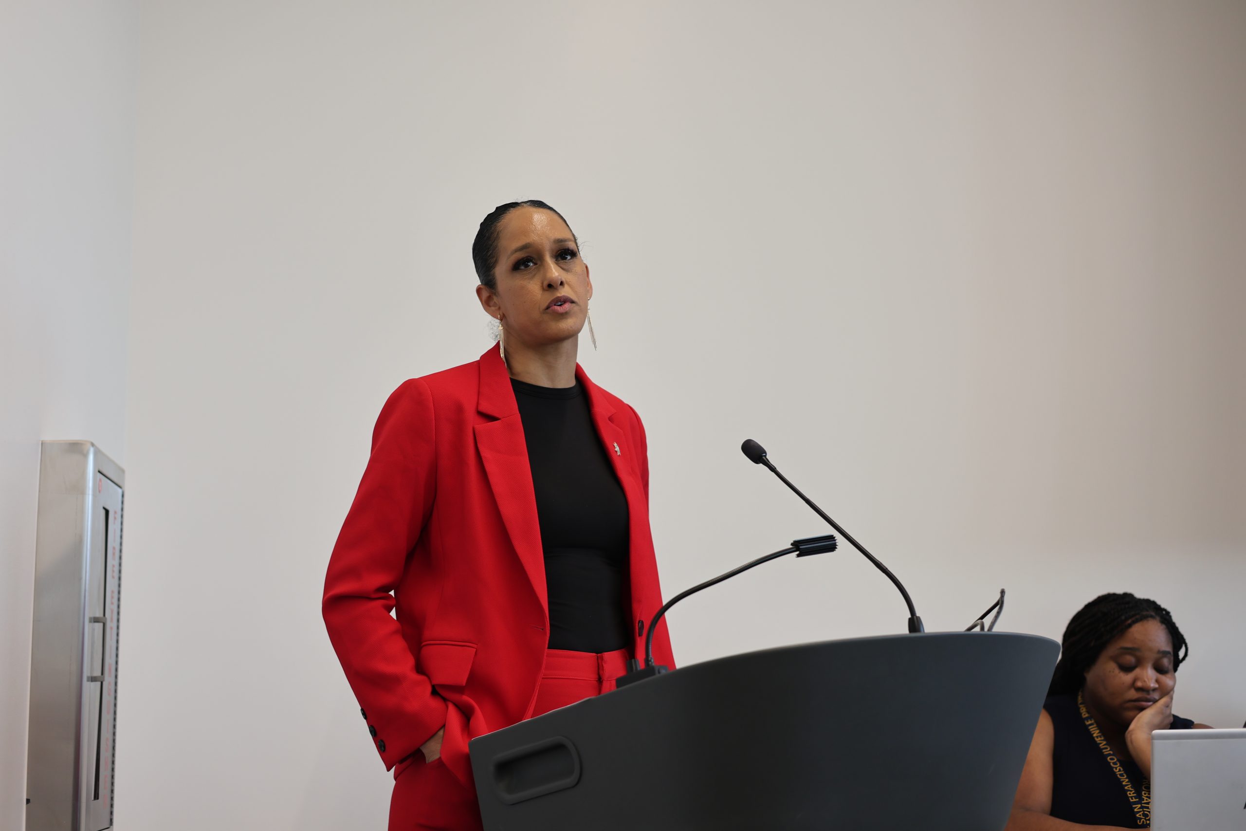 Person in a red suit speaking at a podium with microphones, in front of a plain white background; another person is partially visible seated nearby.