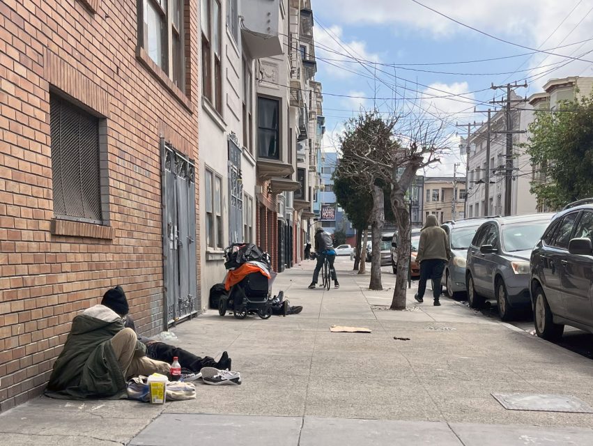 Person sitting on a sidewalk beside a brick building with belongings, a stroller, and food items. Another person is walking away down a street lined with parked cars and residential buildings.