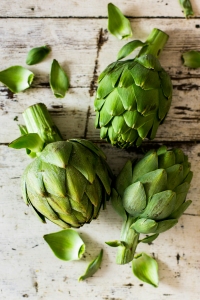 Three green artichokes with scattered leaves on a rustic wooden surface.