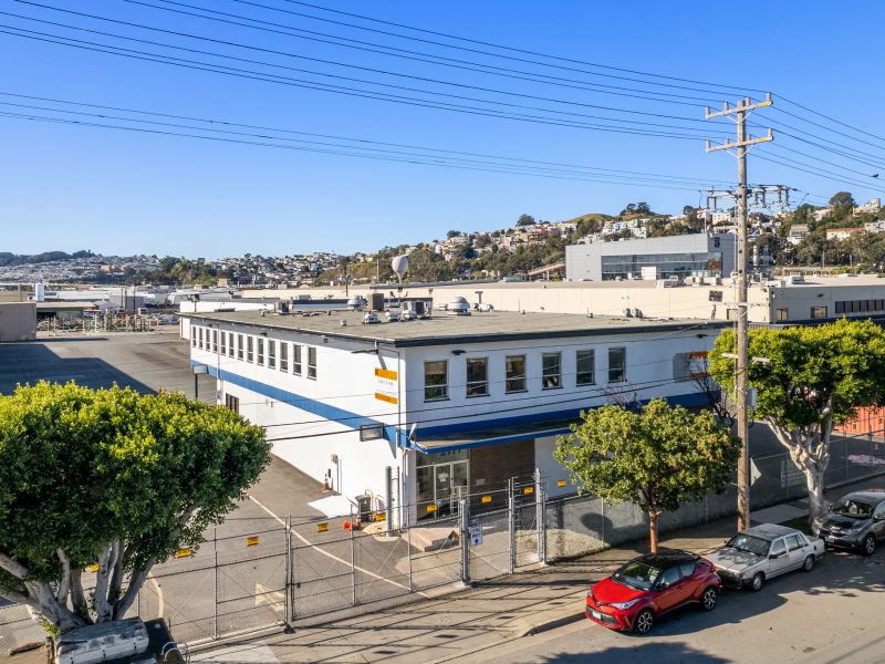 Industrial building with white and blue exterior, located on a street with parked cars and power lines. Surrounded by hills and other industrial structures in the background. Clear blue sky.