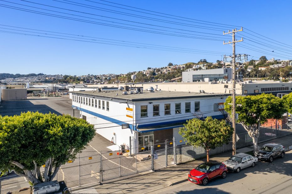 Industrial building with white and blue exterior, located on a street with parked cars and power lines. Surrounded by hills and other industrial structures in the background. Clear blue sky.