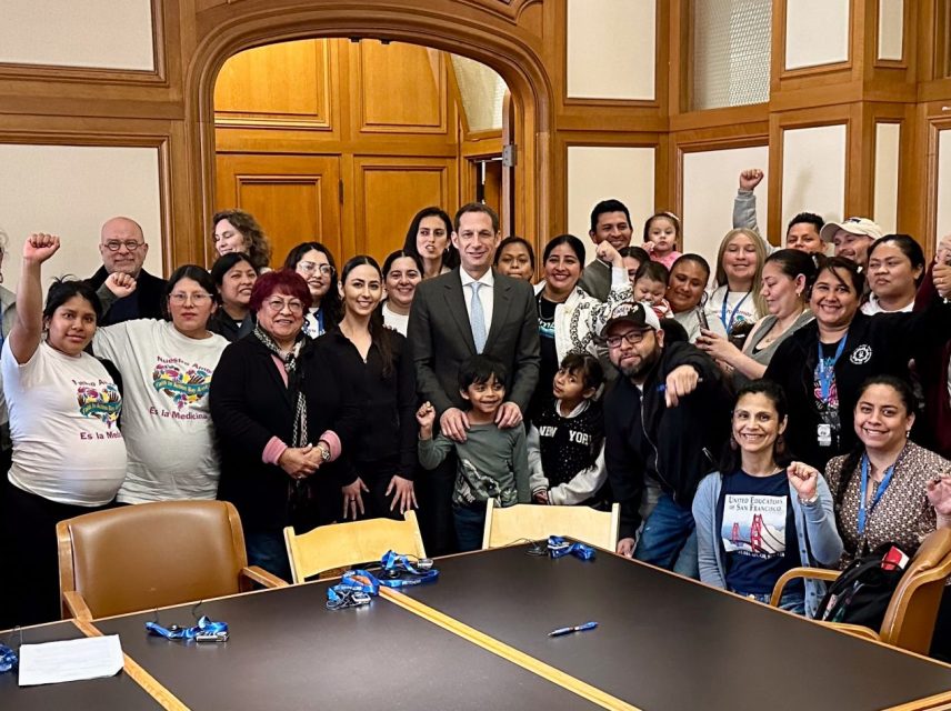 A large group of people gather in a wood-paneled room, some holding up fists. A table with pens is in the foreground.