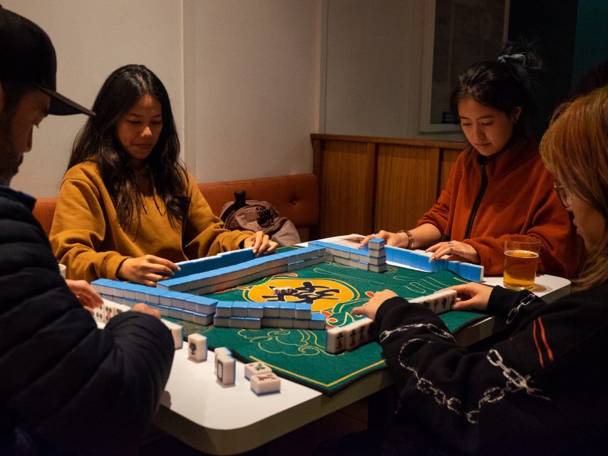Four people playing mahjong at a table with a green mat, concentrating on organizing their tiles.