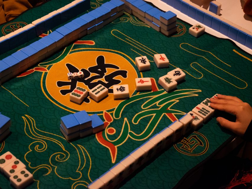 A game of Mahjong in progress with tiles arranged on a green patterned table. A player's hand is visible, with a roll of dice nearby.