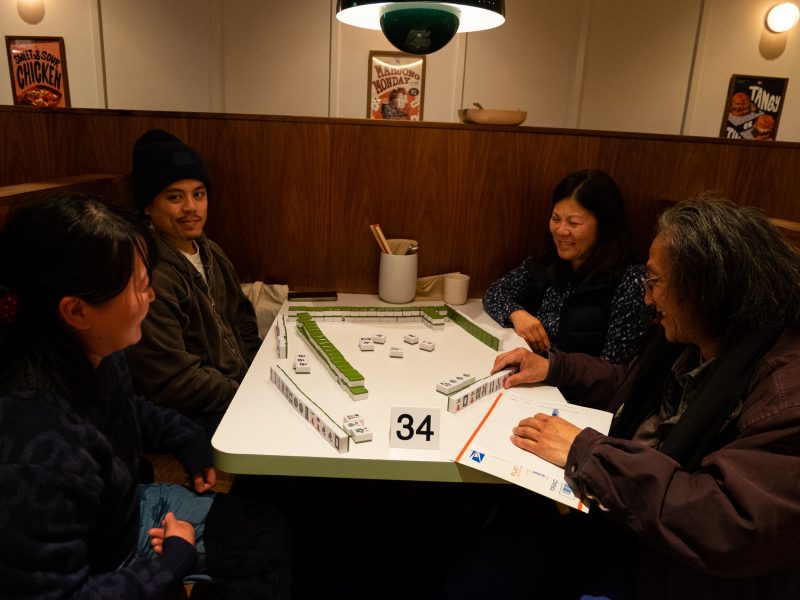 Four people seated in a booth playing dominoes on a table, surrounded by warm lighting and wall posters.